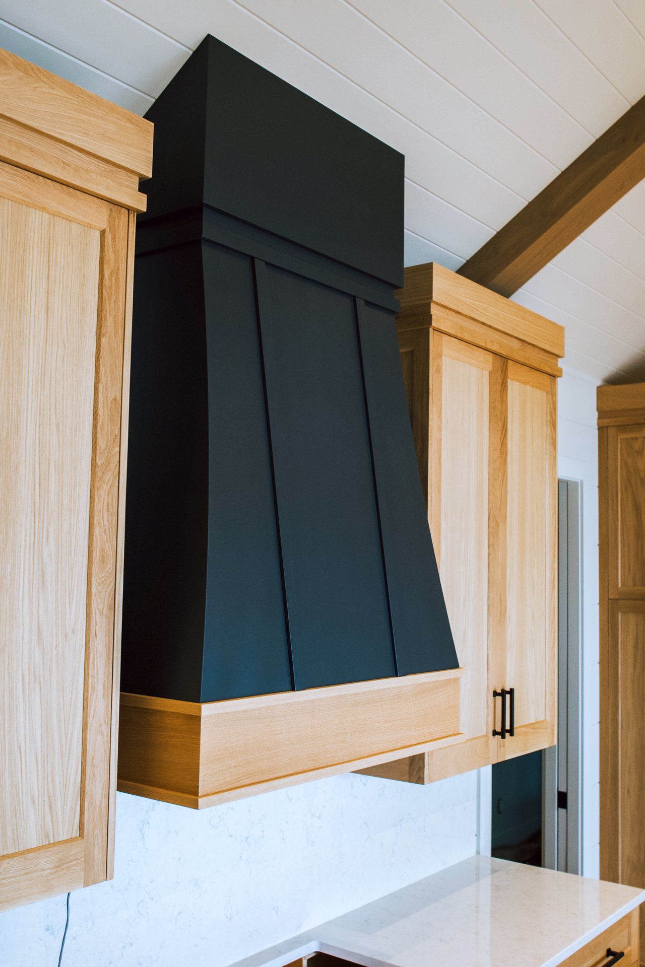 Tapered black range hood with white oak band between white oak upper cabinets